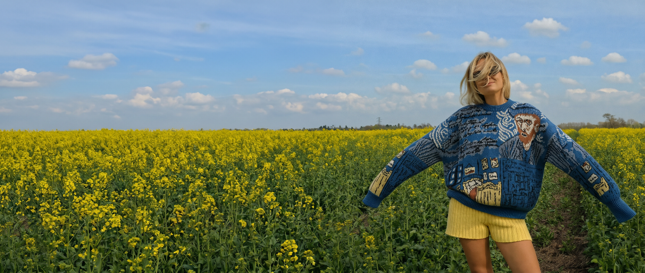 Person wearing a blue sweater with a pattern, standing in a field of yellow flowers under a blue sky.